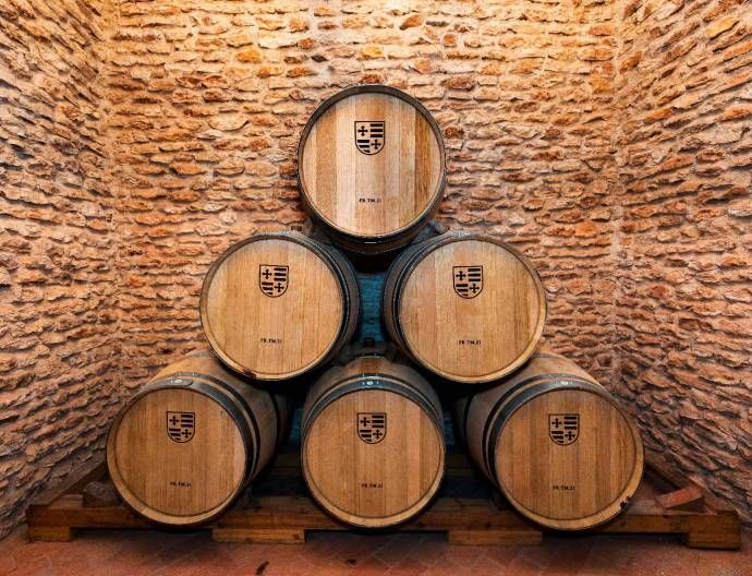 Seis barricas de madera apiladas, dispuestas en forma de pirámide, contra paredes de piedra rústica. Cada barrica lleva estampado un escudo de Bodegas Muñoz