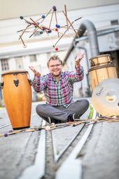 Der Künstler sitzt lachend auf der Straße zwischen Perkussionsinstrumenten und jongliert mit Schlegeln