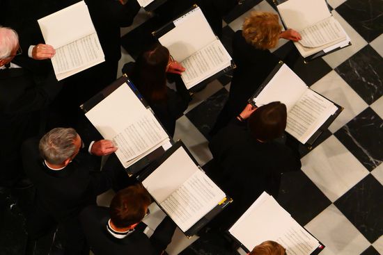 Luftaufnahme von mehreren Chormitgliedern während einer Aufführung. Die Sängerinnen und Sänger in formeller schwarzer Kleidung stehen mit geöffneten Notenmappen in der Hand. Sie stehen in der königlichen Basilika Nuestra Señora de Atocha in Madrid mit ihrem schwarz-weiß gefliesten Fußboden, während sie ihre Noten durchgehen.