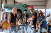 La banda en un taller de coches, instrumentos en mano, un coche antiguo al fondo.